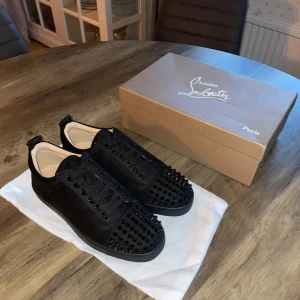 Christian Louboutin med spikar - Svarta sneakers från Christian Louboutin med coola svarta nitar på tån och ovandelen. Skorna har snörning, rund tå och en röd signatursula undertill. Tillverkade i mocka och skinn för en lyxig känsla. Perfekt för dig som vill sticka ut med en edgy stil.