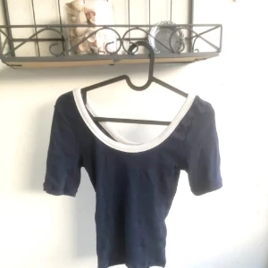 Mörkblå kortärmad topp med vit kant - En enkel och stilren basic mörkblå topp med korta ärmar och bred, vit kant runt den djupa rundade halsringningen. Toppen är ribbad men det syns inte så mycket. Lite tjockare tyg så den är varm oxå (så du vet)