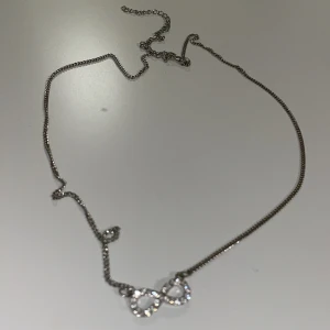 Silverhalsband med hjärtan - Snyggt halsband i silverfärg med två glittriga hjärtan som hängsmycke. Kedjan är tunn och stilren, perfekt för att ge din look lite extra shine. Hjärtana är dekorerade med små stenar som glittrar i ljuset.