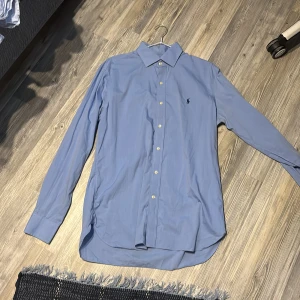 Blå skjorta från Ralph Lauren - Snygg blå skjorta från Ralph Lauren med klassisk krage och knappar framtill. Skjortan har lång ärm och den ikoniska broderade loggan på bröstet. Tillverkad i mjuk bomull som känns skön mot huden. Perfekt för dig som gillar stilrena och tidlösa plagg.