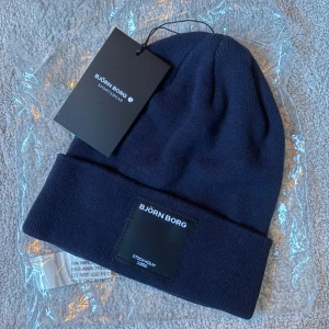Mörkblå Björn Borg-mössa - Helt ny, oanvänd snygg mörkblå mössa från Björn Borg med uppvikt kant och stilren svart patch framtill med logga. Ny med lappar! Perfekt julklapp till den sportiga pojkvännen, flickvännen, brodern, systern, mamman, pappan osv.
