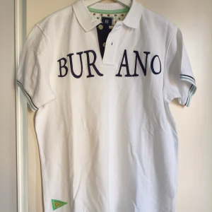 Hansen & Jacob Pikétröja - Vit pikétröja från Hansen & Jacob med Burano broderad text framtill. Klassisk krage, kortärmad modell och gröna detaljer på ärmsluten. Perfekt för en clean och sportig look. Knappslå med två knappar och liten grön patch nertill.Piken är ny utan prislapp.