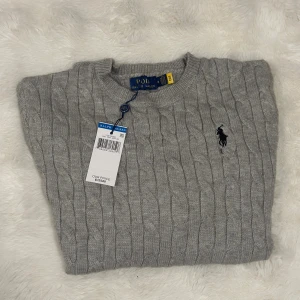 Grå kabelstickad tröja Polo Ralph Lauren - Snygg grå kabelstickad tröja från Polo Ralph Lauren med klassisk rund halsringning och broderad svart logga på bröstet. Tröjan är gjord i mjuk ull och har en tidlös look som passar perfekt till jeans eller chinos.