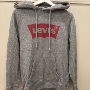 Grå hoodie från Levi's med röd logga - Säljer en grå hoodie från Levi's i storlek XS. Tröjan har en stor röd Levi's-logga framtill, huva med snörning och en klassisk känguruficka. 