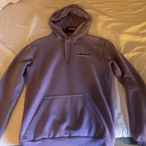 Ljuslila hoodie från Adidas - Säljer en snygg ljuslila hoodie från Adidas med stor logga och text på ryggen samt mindre logga på bröstet. Tröjan har huva med snörning, magficka och ribbade muddar. Perfekt för chill eller streetwear. Använd 2 gånger så de är som ny.