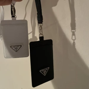  plånbok i svart och vitt - Två stilrena plånböcker, en i svart och en i vitt, med klassisk metalltriangel-logga framtill. Båda har praktisk rem och fack för kort eller ID. Perfekt för att hålla koll på dina viktigaste grejer med lyxig touch.