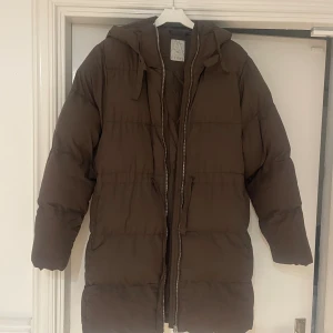 Brun pufferjacka från Etirel - Säljer en brun pufferjacka från Etirel i storlek 34. Jackan har huva med snörning, långa ärmar och dragkedja framtill. Perfekt för kalla dagar med sin långa och fluffiga design. Fickor på sidorna.