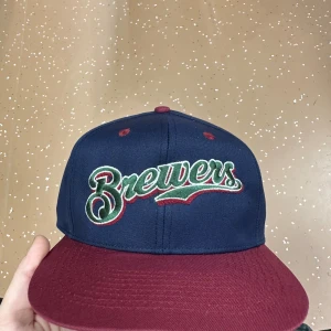 Brewers snapback keps marinblå vinröd - Snygg snapback keps från Brewers med marinblå krona och vinröd skärm. Broderad logga i grönt, vitt och rött framtill. Justerbar baktill med plastspänne. Undersidan av skärmen är grön. Perfekt för dig som vill sticka ut med sportig stil.