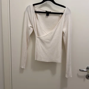 Vit omlottopp från Lindex - Säljer en stilren vit omlottopp från Lindex i ribbad bomullsmix. Toppen har lång ärm, djup v-ringning och sitter snyggt figurnära. Perfekt att matcha med jeans eller kjol för en clean look. Säljer likadan i grönt.