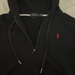 Svart hoodie från Polo Ralph Lauren XL - Svart hoodie från Polo Ralph Lauren i storlek XL med dragkedja och snörning i huvan. Klassisk röd logga broderad på bröstet och custom fit-passform. Perfekt för en chill och stilren look.