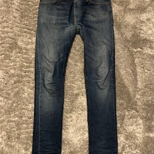 Nudie Jeans - Snygga blå jeans från Nudie Jeans med klassisk femficksdesign och orangea sömmar. Jeansen har en mörk tvätt med lätt slitning och Nudie Jeans-logga på bakfickan och linningen. Perfekt passform för dig som gillar smalare ben.