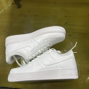 Nike Air Force 1 helvita sneakers - Nike Air Force 1 sneakers i helvit färg med klassisk rund tå och platt sula. Skorna har snörning, perforerade detaljer på tån och är tillverkade i skinn med textilfoder. Snyggt broderad Nike-logga på hälen och ikonisk Air-sula för extra komfort.
