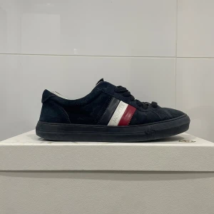 Moncler sneakers i mörkblå mocka - Snygga sneakers från Moncler i mörkblå mocka med klassiska röd, vit och marinblå ränder på sidan. Skorna har rund tå, platt sula och svart snörning. Moncler-logga på plösen och insidan är vit. Perfekt för dig som gillar exklusiva streetwear-vibbar. Stlr 41 passar 42.