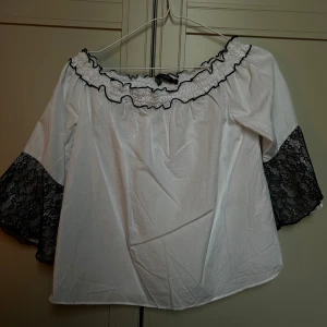 Vit offshoulder blus från Zara - Säljer en vit offshoulder blus från Zara med svarta spetsdetaljer på de vida trekvartsärmarna. Blusen har en rynkad och volangprydd halsringning med svarta kanter som ger en snygg kontrast. 🌟🤍