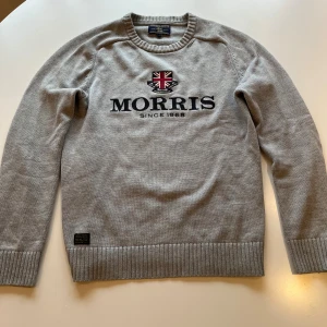 Grå stickad tröja från Morris - Snygg grå stickad tröja från Morris med broderad logga och Union Jack-märke på bröstet. Tröjan har rund hals, ribbade muddar och en klassisk passform. Perfekt till jeans för en stilren look. Tvär fin tröja köpt för några årsedan! Hör av dig vid minsta fundering Mvh Leo.