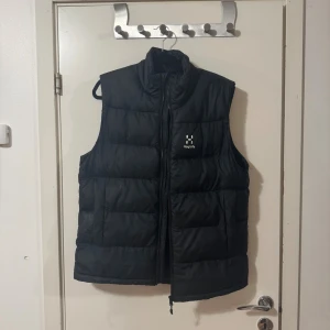 Svart dunväst från Haglöfs - Svart dunväst från Haglöfs med hög krage och quiltad design. Västen har dragkedja framtill och diskret logga på bröstet. Perfekt för lager-på-lager och håller dig varm under kyliga dagar. Ganska varm också höst/vår väst 700kr nypris 2500kr