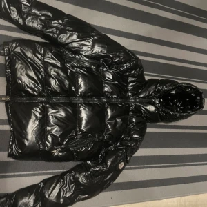 Svart Moncler pufferjacka med huva - Säljer en svart, glansig pufferjacka från Moncler med huva och dragkedja framtill. Jackan har Moncler-logga på ärmen och coolt serietryck på insidan. Perfekt för dig som vill ha en varm och snygg jacka med streetkänsla.