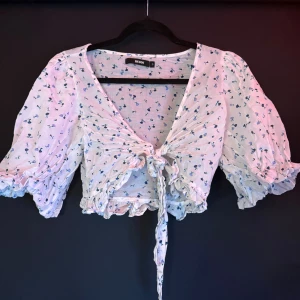 Blommig omlottopp från Bik Bok - Supersöt vit omlottopp från Bik Bok med blått blommönster. Toppen har puffiga korta ärmar, v-ringning och knyt framtill. Croppad modell med volangkant nedtill, perfekt för vår och sommar. Tunn och lätt bomullskänsla.