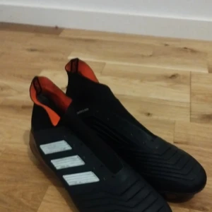 Svarta adidas fotbollsskor med orange detaljer - Säljer ett par svarta adidas fotbollsskor med vita ränder på sidan och orange insida. Skorna har en strömlinjeformad design utan snörning och mönstrad ovansida för bättre bollkontroll. Yttersulan har dobbar i svart och brons för bra grepp på planen.