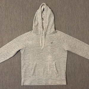 Ljusgrå hoodie från Hollister XS - Mysig ljusgrå hoodie från Hollister i fleece med dragsko i huvan och känguruficka framtill. Tröjan har en liten Hollister-logga på bröstet och är perfekt för chill dagar. Långärmad och i mjukt material som känns skönt mot huden.