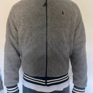 Grå teddyjacka från Polo Ralph Lauren - Mjuk och fluffig grå teddyjacka från Polo Ralph Lauren med hög krage och hel dragkedja. Jackan har långa ärmar och ribbade muddar med vita och marinblå ränder. Liten Polo-logga broderad på bröstet. Perfekt för kyliga dagar.