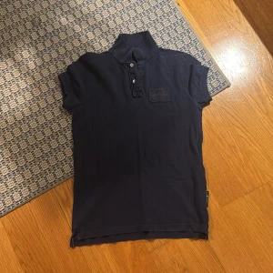 Marinblå pikétröja Sail Racing S - Snygg marinblå pikétröja från Sail Racing i storlek S. Klassisk krage, korta ärmar och knappslå framtill. Diskret logga på bröstet och mjukt bomullsmaterial som känns skönt mot huden. Perfekt för dig som gillar sportig och stilren look.