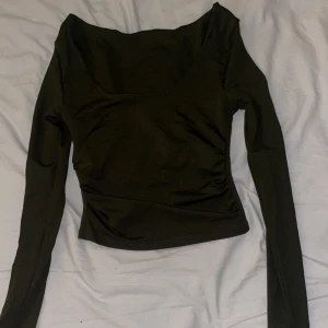 Mörk grön långärmad topp med bred hals - Snygg mörk grön långärmad topp med bred, djup halsringning och figurnära passform. Materialet känns mjukt och stretchigt, perfekt för att skapa en clean och stilren look. Toppen är enkel utan mönster eller detaljer, vilket gör den lätt att matcha med olika outfits.