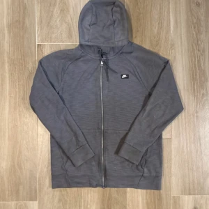 Grå hoodie från Nike med dragkedja - Säljer en grå hoodie från Nike med hel dragkedja och huva med snörning. Tröjan har långärmade raglanärmar och en liten Nike-logga på bröstet. Perfekt för chill eller träning, enkel och stilren design.