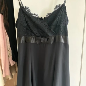 Svart klänning med spets och rosett - Svart klänning med tunna axelband, spets upptill och en söt rosett i midjan. Överdelen har blommig spets och en satinliknande kant. Perfekt för dig som gillar eleganta detaljer och stilren look.