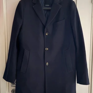 Mörkblå rock från J.Lindeberg - Snygg mörkblå rock från J.Lindeberg med klassisk krage och tre knappar framtill. Jackan har två sidofickor och en bröstficka. Yttermaterialet är 80% ull och 20% polyamid, fodret är 100% viskos. Perfekt för dig som gillar stilrena och tidlösa plagg.