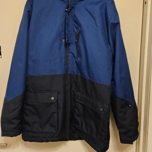 Blå och svartskid/ vinter jacka från Ski Industries - Säljer en blå och svart skid jacka från Ski Industries, storlek L. Jackan har huva, dragkedja framtill, stora fickor med knappar och en bröstficka med dragkedja. Materialet är polyester och jackan är vind- och vattenavvisande. Perfekt för kalla dagar och outdoor. Använd 3 dagar i fjällen. 