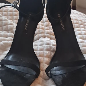 Svarta klackade sandaler från Even&Odd - Svarta sandaler med smal rem över tårna och justerbar ankelrem med guldfärgat spänne. Klackarna är höga och smala, vilket ger en elegant siluett. Skorna har öppen tå och är tillverkade i mocka-liknande material för en mjuk känsla.