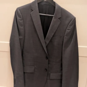 Mörkgrå kavaj från Hugo - Snygg mörkgrå kavaj från Hugo med två knappar framtill och klassiska fickor med lock. Insidan har coola röda detaljer och innerfickor. Perfekt för dig som vill ha en stilren och modern look. Materialet känns lyxigt och är troligtvis ullblandning. Original priset 2500kr