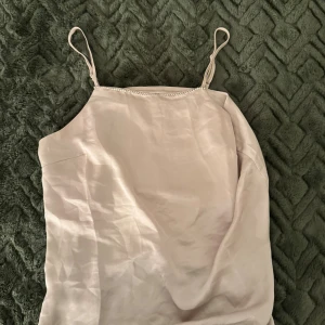 Champagnefärgad linnetopp med strass - Champagnefärgad linnetopp från Kappahl med tunna axelband och glittrande strassdetaljer längs halsringningen. Toppens material har en silkeslen finish och en lätt, flowy passform som känns lyxig. Perfekt för dig som gillar stilrena och glittriga detaljer.