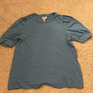 Blå topp med puffärm från Vero Moda - Säljer en ljusblå topp från AWARE by Vero Moda i storlek XS. Toppen har puffiga korta ärmar med breda muddar och rund halsringning. Materialet är mjuk bomull.