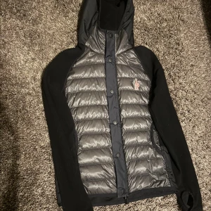 Svart och grå dunjacka från Moncler - Snygg dunjacka från Moncler i svart och grått med quiltad framsida och huva. Jackan har Moncler-logga på bröstet, dragkedja och knappar framtill. Ärmarna är svarta och materialet är en mix av polyester och dun, perfekt för kyliga dagar.