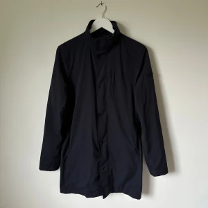 Mörkblå rock från J.Lindeberg, storlek S - Mörkblå rock från J.Lindeberg med stilren design och hög krage. Jackan har knäppning framtill och en diskret bröstficka. Tillverkad i 100% polyester med foder i polyamid, vilket gör den lätt och smidig. Perfekt för dig som gillar minimalistisk och clean look. Modell: Terry 72 Tech Stretch - 4D6