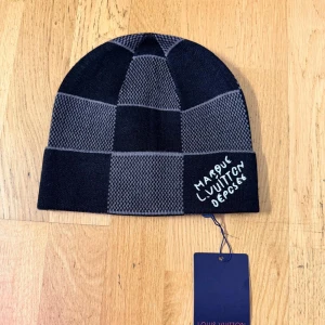 Loui buitton rutig svart/grå mössa - Exklusiv mössa från Loui buitton i svart och grått med rutigt mönster. Broderad text 'Marque L. Vuitton Deposee' framtill. Klassisk beanie-modell i mjukt stickat material, perfekt för att lyfta din vinterstil.