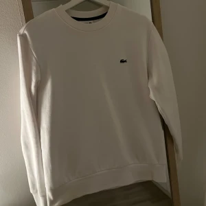 Lacoste Sweatshirt  - Vit sweatshirt från Lacoste i Classic Fit med den ikoniska gröna krokodilen broderad på bröstet. Tröjan har rund hals, långa ärmar och ribbade muddar. Perfekt för dig som gillar stilrena och klassiska plagg med snygg logga. 