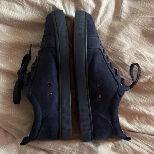  Christian Louboutin  - Snygga mörkblå sneakers från Christian Louboutin i mocka med klassisk röd sula och rund tå. Skorna har snörning och diskreta metalldetaljer vid snörhålen. Insidan är ljusbeige och sulan har Louboutin-loggan i rött. Perfekt för dig som gillar exklusiva märken och stilren design. Skolåda, kvitto o annat följs med (bild 2)