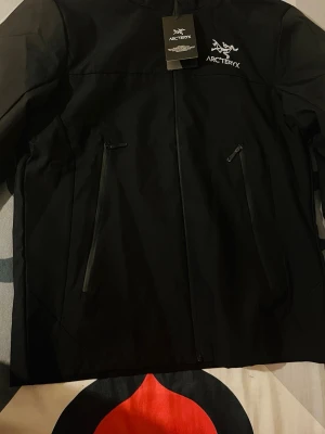 Svart vindjacka från Arc'teryx - Snygg svart vindjacka från Arc'teryx med dragkedja framtill och två stora fickor med dragkedja. Jackan har broderad logga på bröstet och är tillverkad i ett slitstarkt, lätt material som står emot vind. Perfekt för dig som vill ha en stilren och funktionell jacka.