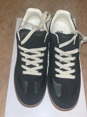 Svarta sneakers med vit sula - Stilrena svarta sneakers med vita snören och kontrasterande vit insida. Skorna har en klassisk låg profil och en brun gummisula som ger en retrovibe. Materialet ser ut att vara en mix av läder och mocka för en snygg och tidlös look.