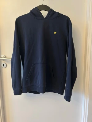 Lyle and scott hoodie  - Snygg mörkblå hoodie med huva och magficka. På bröstet sitter en liten gul broderad logga. Hoodien har en klassisk passform och är tillverkad i mjukt material som känns skönt mot huden. Perfekt för en avslappnad och stilren look. Notera även skador på armarna!