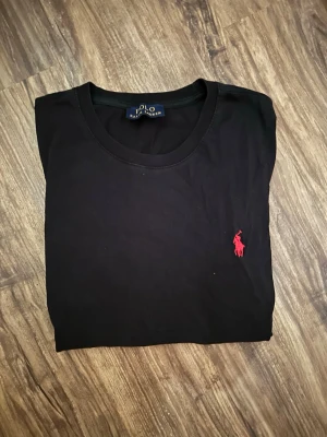 Svart t-shirt från Polo Ralph Lauren - Snygg svart t-shirt från Polo Ralph Lauren med klassisk röd logga broderad på bröstet. T-shirten har rund halsringning och korta ärmar. Tillverkad i mjukt bomullsmaterial som känns skönt mot huden. Perfekt basplagg för en clean och stilren look.