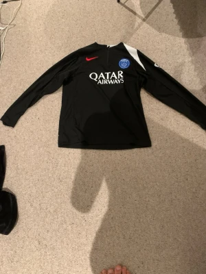 Svart PSG träningströja långärmad - Säljer en svart långärmad träningströja från PSG med vit och röd detalj, halv dragkedja och tryckta loggor på bröstet och ryggen. Materialet är troligtvis polyester, perfekt för fotbollsträning eller sport. Passar 13 till 14 åringar