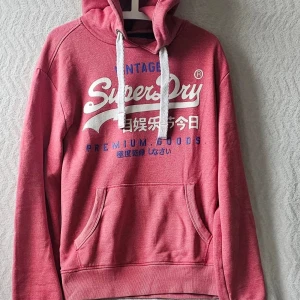 Rosa hoodie från Superdry med tryck - Säljer en rosa hoodie från Superdry med vit och blå text framtill. Hoodien har huva med vita snören och en stor magficka. Mjuk insida i fleece och klassisk passform. Perfekt för dig som gillar streetwear och vill ha något bekvämt och snyggt.