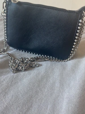 Svart axelväska med kedja och nitar - Snygg svart axelväska i slätt konstläder med silverfärgade kedjedetaljer och nitar längs kanterna. Väskan har en lång kedjerem och stängs med dragkedja upptill. Perfekt för dig som gillar en edgy och trendig look.