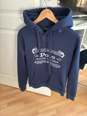 Marinblå hoodie från Polo Ralph Lauren - Snygg marinblå hoodie från Polo Ralph Lauren med vit logga och text på bröstet. Klassisk känguruficka framtill och justerbar huva med dragsko. Perfekt för en avslappnad och trendig stil.