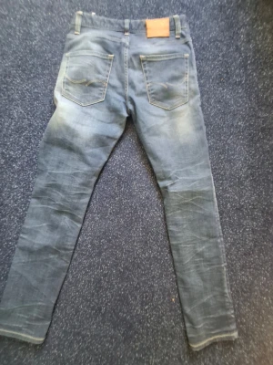 Blå slim fit jeans med slitningar - Säljer ett par blå slim fit jeans med snygga slitningar och tvättade detaljer. Jeansen har två bakfickor med dekorativa sömmar och en klassisk läderpatch i midjan. Perfekta för dig som gillar en avslappnad men trendig stil. Det står inte men jag skulle tro 29 32