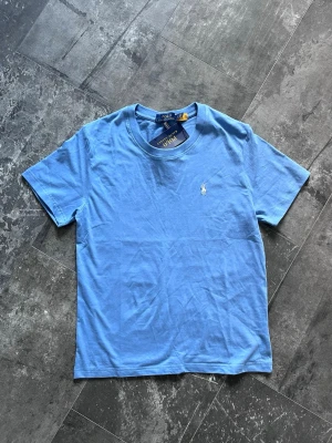 Blå t-shirt från Polo Ralph Lauren - Snygg ljusblå t-shirt från Polo Ralph Lauren med klassisk passform och korta ärmar. T-shirten har en liten broderad logga på bröstet och är gjord i mjuk bomull. Perfekt till jeans eller shorts för en clean look.